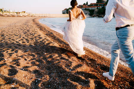 Bride and groom run along the sandy shore against the backdrop of Sveti Stefan islandの写真素材
