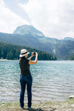 Woman stands on the shore and takes pictures of the mountains on her phone. Back view