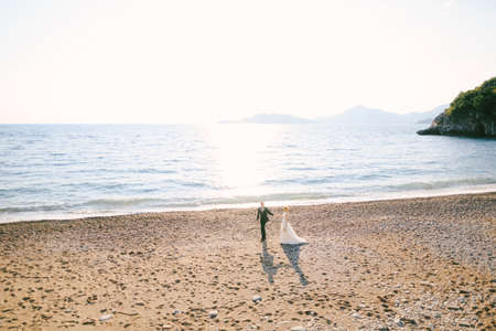 Groom and bride in a white dress with a bouquet of flowers holding hands are walking along the beach by the sea against the background of green mountainsの写真素材
