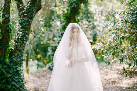Bride in a white lace dress, covered with a veil, stands among the ivy-covered trees
