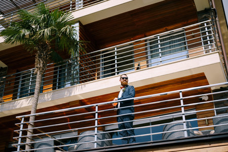 Groom stands on the balcony of the hotel with a glass in his hand leaning on a metal fenceの写真素材