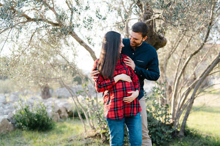 Smiling man hugging shoulders of pregnant woman from behind in parkの写真素材