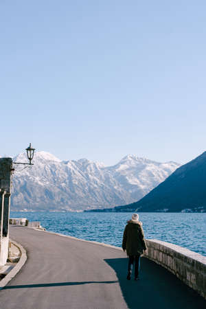 Girl in a jacket walks along the asphalt embankment against the background of the sea and mountainsの写真素材