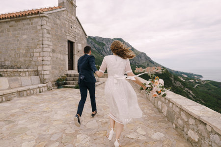 Groom and bride with a bouquet of flowers run to the church on the mountain. Back viewの写真素材