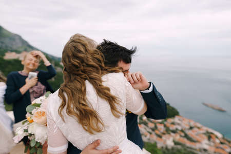 Groom hugs bride with a bouquet of flowers while standing on the mountain. Back viewの写真素材