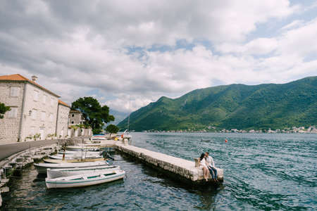 Man and woman are sitting on the pier by the water next to the moored boatsの写真素材