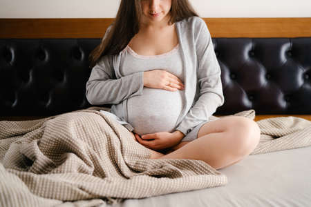 Pregnant woman hugs belly with hands while sitting on bedの写真素材