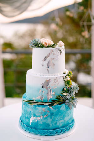 White and blue wedding cake with flowers and succulents stands on a cake-stand on a white tableの写真素材