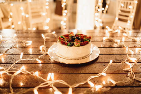 White wedding cake with fruit on top on a wooden table decorated with garlands of light bulbsの写真素材