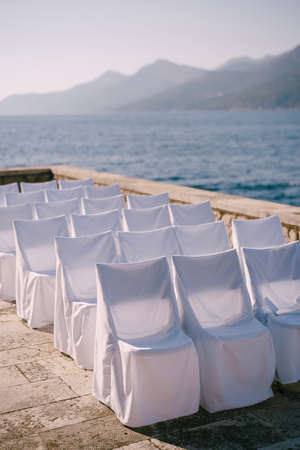 Row of chairs in white fabric covers stands near a stone fence on the seashoreの写真素材