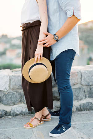 Man hugs woman from behind while standing by a stone fence against a background of mountainsの写真素材