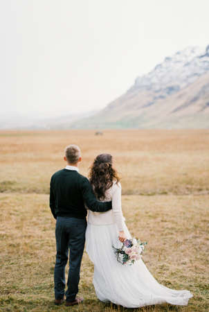 Groom hugs bride with a bouquet on a dry meadow at the foot of the mountains. Back viewの写真素材