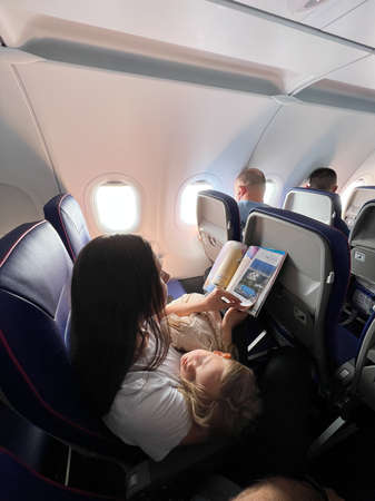 Mom with a sleeping daughter in her arms sits in an airplane seat and reads a magazineの写真素材