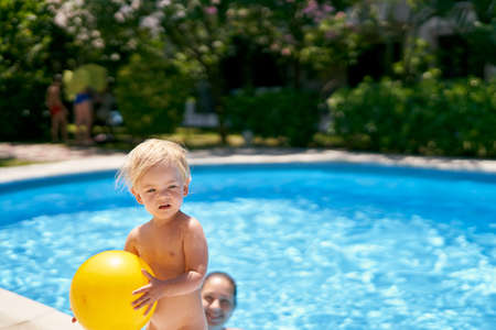 Small baby with a yellow ball stands by the pool with turquoise waterの写真素材