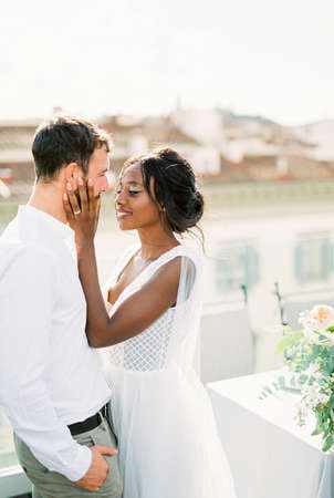 Smiling bride touches groom face with hands on terrace at homeの写真素材