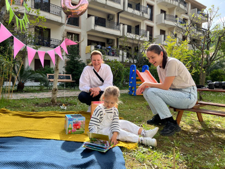 Budva, Montenegro - 07.07.22: Little girl sits on a blanket and leafs through a book next to her mother and grandmotherのeditorial素材