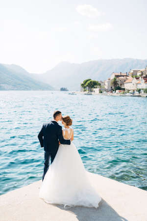 The bride and groom are standing, hugging and kissing on the pier near the old town of Perast, back viewの写真素材