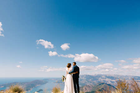 Newlyweds stand with their backs hugged against the backdrop of mountains and a bright blue skyの写真素材