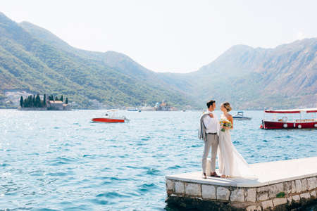The bride and groom standing side by side on the pier in the Bay of Kotor, islands of Perast are behind themの写真素材