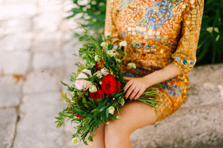 A bride in an unusual golden dress with icon sits and holds a bridal bouquet of white and red roses in her hands, close-upの写真素材