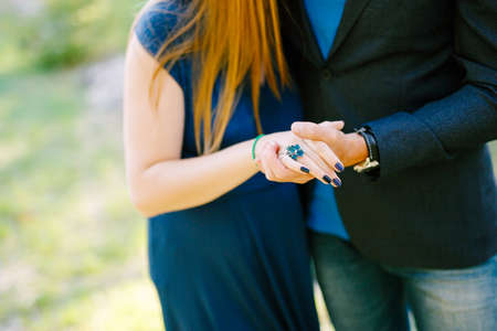 Man in a jacket holds the hand of woman in a long dress with a blue ring on her finger and a...