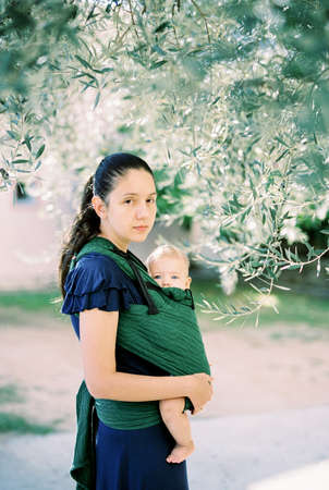 Mom with a baby in a sling stands in an olive grove under a treeの写真素材