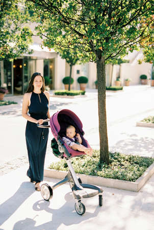 Mom in a long dress stands with a stroller with a baby under a tree