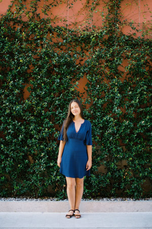 Young woman in a blue dress stands near a wall covered with green ivyの写真素材