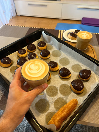 Glass of cappuccino in a man hand over a baking sheet with custard cakes with craquelins