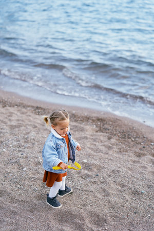 Little girl in a dress and a blue denim jacket carries sand in a toy rake on the beach near the...