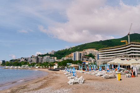 Budva, Montenegro - 16.08.2022: Sun loungers with folded umbrellas stand on the beach against the backdrop of buildings, greenery and mountainsのeditorial素材