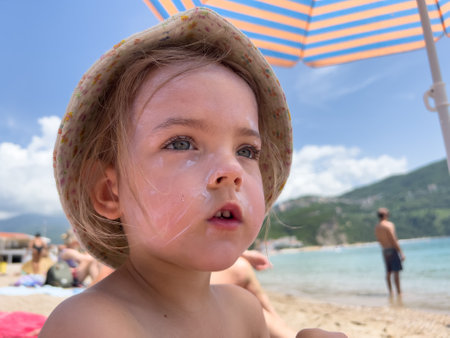 Little girl with sunscreen on her face sits on the beach under sun umbrellaの写真素材