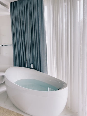 Oval bathtub with chrome taps stands near a panoramic window with curtainsの写真素材