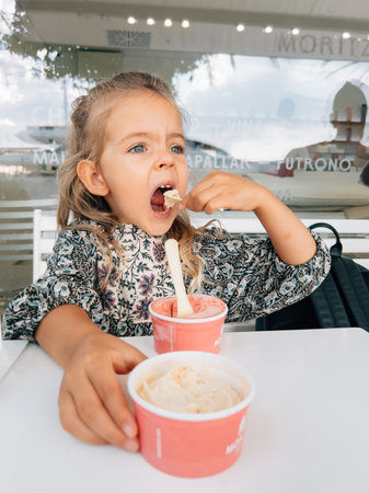 Little girl eating ice cream from two cups with a spatula at the tableの写真素材