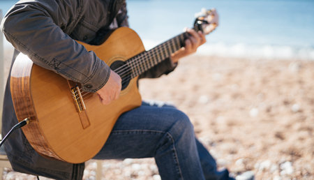 Musician plays the acoustic guitar while sitting on a chair on the beach. Cropped. Faceless