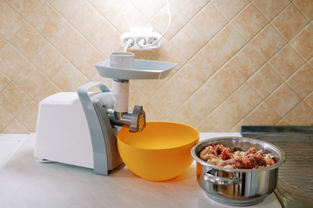 Electric meat grinder stands on a table next to a bowl and a pot of minced meat