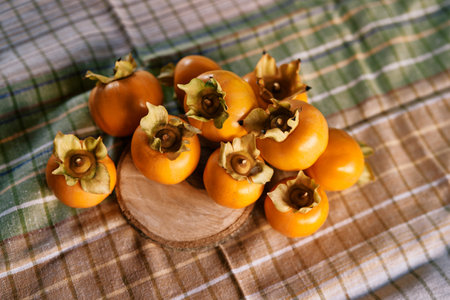 Pile of ripe persimmon lies on a checkered tablecloth and a wooden round board on the table. Top...