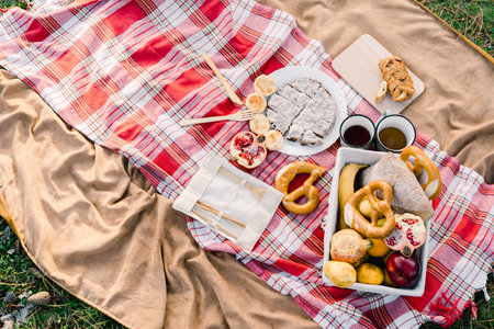 Basket with pastries and fruits stands next to mugs of coffee on a blanket on green grassの写真素材