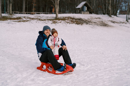 Smiling dad with little daughter are sitting on a sled on a snowy plainの写真素材
