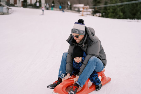 Smiling dad with a little boy riding down a snowy hill on a sledの写真素材