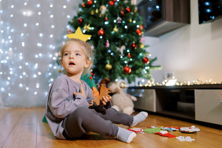 Little girl sits near the Christmas tree on the floor with felt toys in her hands