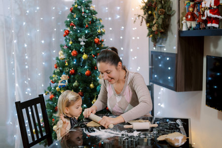 Mom rolls out a piece of dough to a little girl with a rolling pin while sitting at the table with herの写真素材
