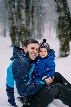Smiling dad with a little girl on his knees sits under a snowfall in the forestの写真素材