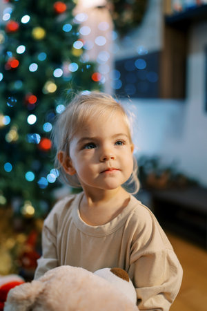 Little girl sits near a Christmas tree sparkling with colorful lights. Blur