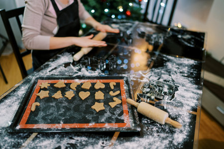 Cook rolls out the dough on the table next to the tray with cut out cookies. Cropped