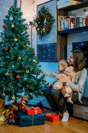 Little girl hangs a ball on a Christmas tree while sitting on her mother lapの写真素材