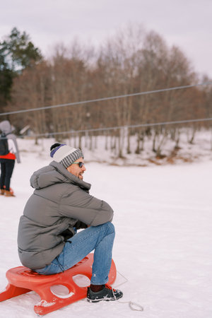 Man in sunglasses sits on the snow and looks away. Side view