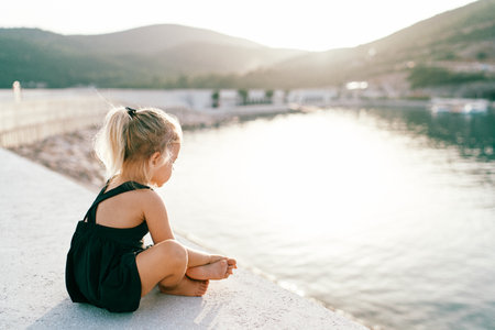 Little barefoot girl sits cross-legged on the pier and looks at the water. Side viewの写真素材
