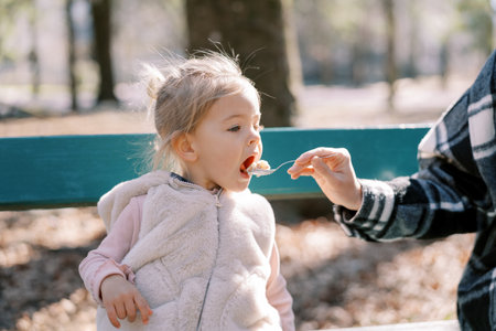 Mom feeds porridge from a spoon to a little girl sitting on a bench in the park. Croppedの写真素材