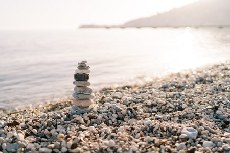 Cairn stands on a pebbly beach near a sparkling sea against the backdrop of mountainsの写真素材